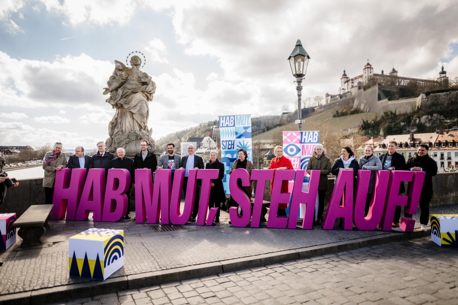 Menschen halten auf Brücke in Würzburg große, pinke Buchstaben, die den Schriftzug „HAB MUT. STEH AUF!“ bilden.